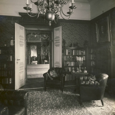 czarno-białe zdjęcie przedstwia wnętrze pokoju, gdzie na podłodze leży dywan po prawej znajdują się fotele, stolik oraz regały z książkami.