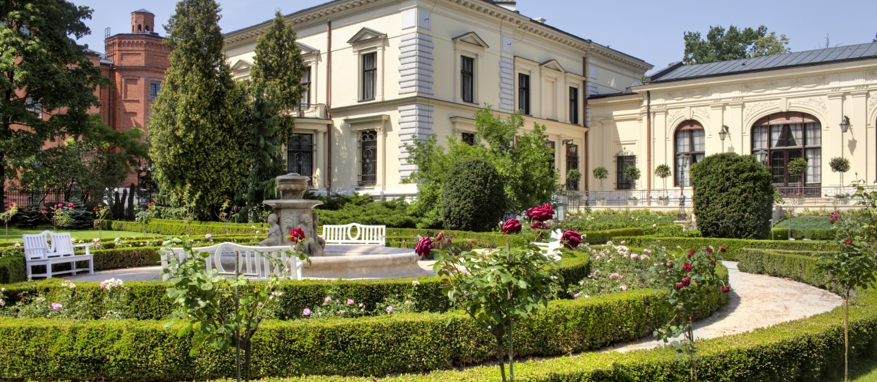 A bright, multi-story palace with a French garden, low hedges, and a fountain in the foreground.