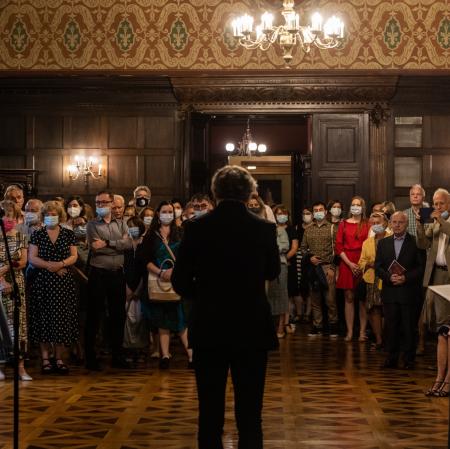 Na pierwszym planie trzy osoby stojące tyłem. W głębi kilkadziesiąt osób zwróconych do przodu.