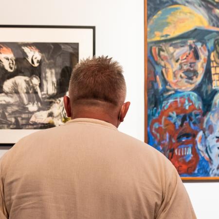 Na pierwszym planie odwrócony tyłem mężczyzna. Na drugim planie dwa obrazy