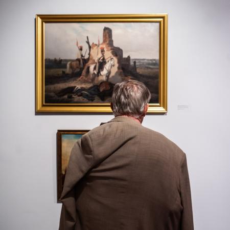 Odwrócony tyłem mężczyzna. Stoi przed ścianą, na której wiszą - jeden nad drugim - dwa obrazy