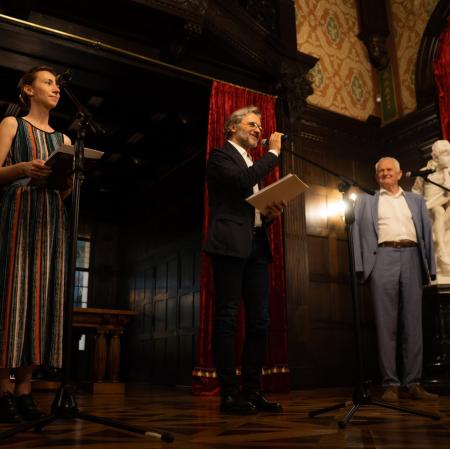 Zdjęcie wykonane z żabiej perspektywy. Przedstawia trzy osoby stojące przy mikrofonach.