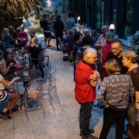 Goście w ogrodzie zimowym. Niektórzy siedzą przy okrągłych stolikach. Inni rozmawiają stojąc w kilkuosobowych grupkach.