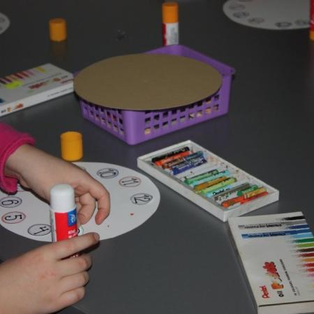 Zbliżenie na ręce dziewczynki przyklejającej  cyfry do okrągłego zegara. Na stole pastele, klej.