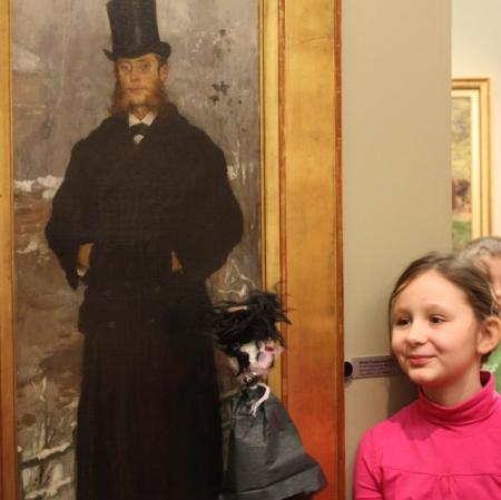 Uczestniczka warsztatów z kukiełką przed obrazem "Portret Kazimierza Bartoszewicza" Witolda Pruszkowskiego.