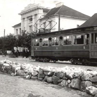 Czarno-białe zdjęcie przestawia tramwaj w zabudowie miejskiej.