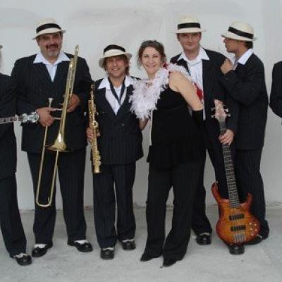 Zdjęcie przedstawia siedem osób. Kilka z nich trzyma instrumenty muzyczne. W środku stoi kobieta. Muzycy ubrani są w czarne stroje. Mężczyźni mają na głowach jasne kapelusze z czarną lamówką. 