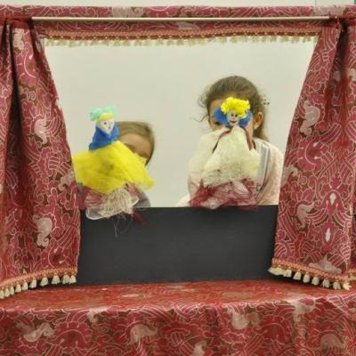 Dwie dziewczynki z kukiełkami wyglądają przez okienko małego teatrzyku. 