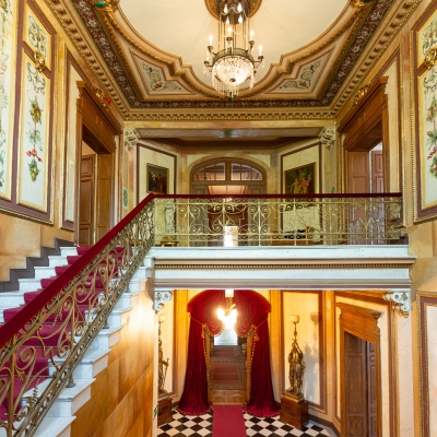 An opulent two-story grand foyer featuring a marble staircase with a red carpet runner, ornate gold railings, checkered floor tiles, and elaborate ceiling moldings surrounding a central crystal chandelier.