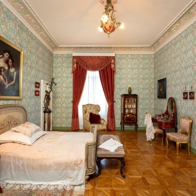 Elegancka sypialnia w stylu retro z zieloną, wzorzystą tapetą i pałacowym parkietem. Po lewej stronie stoi zasłane łóżko z ozdobnym wezgłowiem, nad nim wisi duży portret w złotej ramie, a w głębi widać okno z czerwonymi zasłonami oraz toaletkę z lustrem.