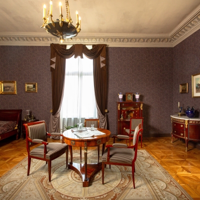 A classic, dark-walled bedroom featuring a sleigh bed, dark brown drapes, a round wooden table with matching chairs, vintage furniture, and a large patterned floor rug.