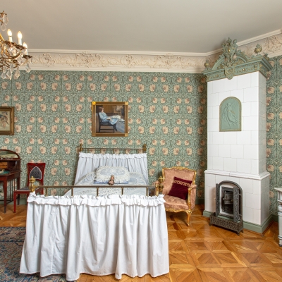 A charming, vintage maiden's bedroom featuring floral patterned wallpaper, a brass bed with white ruffles, a tall white tiled stove, a writing desk, and a washstand.