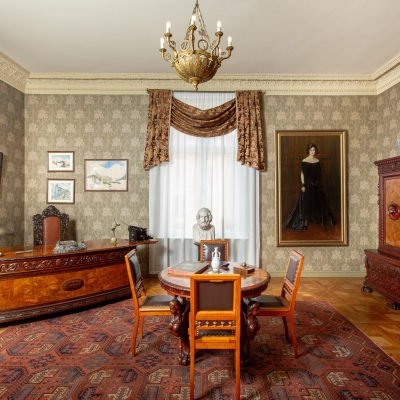 A classic, elegantly furnished study with a large ornate wooden desk, a central round table with chairs, a carved cabinet, a patterned area rug, and framed artwork.