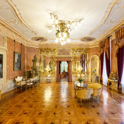 A grand mirror hall featuring polished parquet flooring, gold-framed wall mirrors, ornate stucco ceiling details, and rich red window drapes.