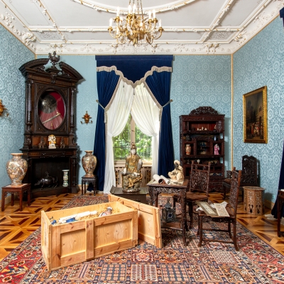 An oriental-style parlor featuring light blue patterned wallpaper, deep blue velvet curtains, ornate dark wood furniture, decorative Asian ceramics, and an open wooden crate on an oriental rug.