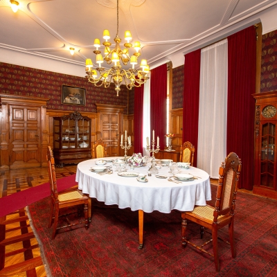 A formal dining room featuring a set oval table with a white tablecloth, high-backed wooden chairs, deep red curtains, dark wood paneling, and a warm, glowing chandelier overhead.