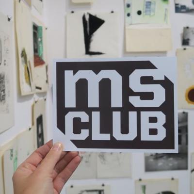 Zdjęcie przedstawia trzymaną w dłoni czarną kartkę z białymi literami: ms club