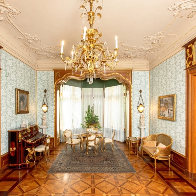 An elegant sitting room featuring light blue floral wallpaper, an ornate gold chandelier, a piano along the side wall, and a small seating group positioned in front of a sunlit alcove window.