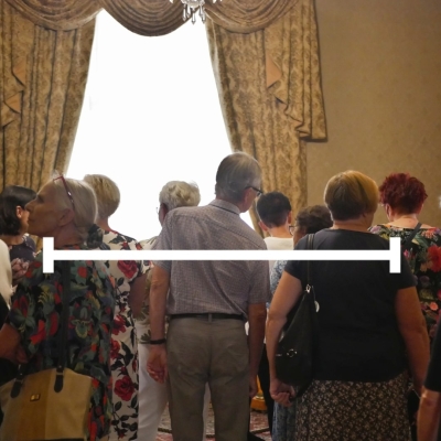 Alternatywny tekst dla tego obrazu brzmi:  Zdjęcie przedstawia grupę dorosłych osób w pomieszczeniu o eleganckim wystroju. W tle widać duże okno z jasnymi zasłonami oraz kryształowy żyrandol. Ludzie stoją zwróceni plecami do widza, wpatrując się w kierunku okna, jakby oglądali coś na zewnątrz lub słuchali kogoś stojącego w tamtej części pomieszczenia. Na pierwszym planie znajduje się mężczyzna w okularach, ubrany w koszulę w kratę, oraz kobieta z jasnymi, krótkimi włosami, w czarnej bluzce. W poprzek obrazu