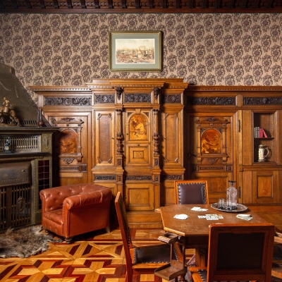 A rustic hunting lodge parlor featuring rich wooden wall paneling, a stone fireplace with leather armchairs, patterned wallpaper, and a card table set with playing cards and chairs.