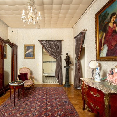Ornate 19th-century bedroom with floral wallpaper, an antique upholstered bed, red window drapes, vintage wooden furniture, and a parquet floor.