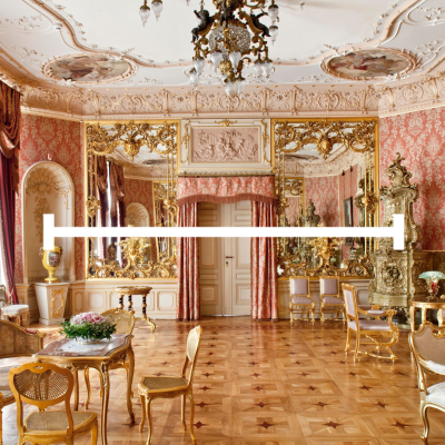 Wnętrze bogato zdobionego, zabytkowego pokoju. Ściany pokrywa tapeta w kolorze łososiowym ze wzorami. Sufit jest ozdobiony sztukaterią i malowidłami. Na środku sufitu wisi duży, ozdobny żyrandol. W pokoju znajdują się meble w stylu rokoko, w tym złocone krzesła i stoliki. Na jednej ze ścian wiszą duże, złocone lustra, a na środku widać dwuskrzydłowe drzwi. W pokoju widać również kominek i zegar.