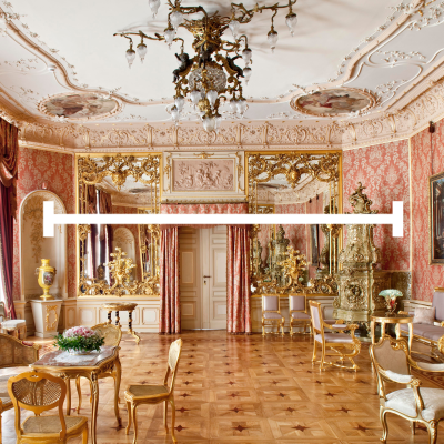 Bogato zdobione wnętrze pałacowe z ozdobnym żyrandolem, wzorzystą tapetą w kolorze łososiowym i kunsztownymi sztukateriami na suficie. Na środku zdjęcia znajduje się duża, biała, pozioma linia zakończona pionowymi kreskami. Pomieszczenie wyposażone jest w złocone meble, liczne lustra w ozdobnych ramach oraz dekoracyjny piec kaflowy po prawej stronie. Podłoga wykonana jest z jasnego parkietu ułożonego w geometryczne wzory.