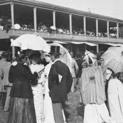 czarno-białe zdjęcie przedstawia kobiety w długich sukniach i mężczyzn w garniturach i cylindrach na głowach. Tłum ludzi dookoła. 