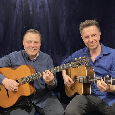 Zdjęcie przedstawia dwóch mężczyzn z gitarami. Mężczyźni uśmiechają się.