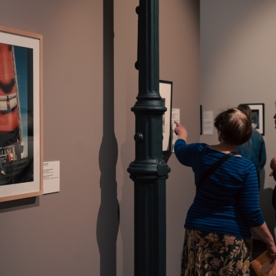 The photo shows the interior of an art gallery or museum. On the right, two women are standing and looking at the exhibition; one of them, wearing a blue striped blouse and a patterned skirt, is pointing at a small artwork hanging on the wall. Next to her stands the other woman in a red blazer and light-colored pants, looking in the same direction. In the foreground on the left, a section of the wall is visible, featuring a large, vertical painting or photograph in a light-colored frame, dominated by a huge