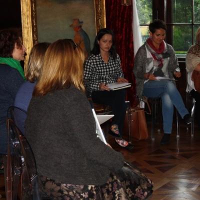 kilka dorosłych siedzi w dużej sali w kręgu obok siebie. Za kobietami obraz na sztaludze