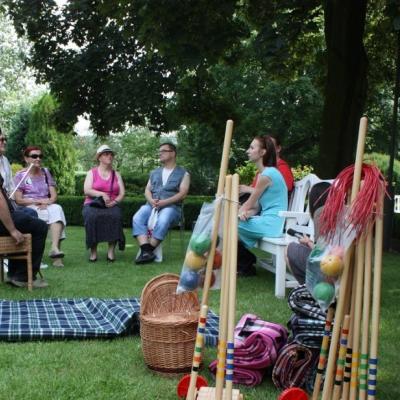  wśród zielonych drzew na trawie drewniane młoteczki, kolorowe kule, koce. Na drugim planie kilka osób dorosłych siedzi na krzesłach w kręgu