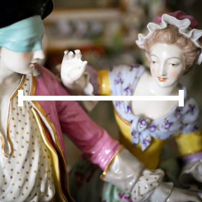 Obraz przedstawia zbliżenie na porcelanowe figurki w stylu rokokowym. Figurka kobiety ubrana jest w bogato zdobioną suknię w pastelowych kolorach, z żółtymi i fioletowymi elementami oraz koronkowym czepkiem na głowie. Druga figurka, przedstawiająca mężczyznę, ma na sobie różową marynarkę, białą koszulę w złote kropki i błękitną maskę zasłaniającą oczy. Kobieta unosi rękę w kierunku mężczyzny. Tło jest rozmyte, ukazując więcej porcelanowych dekoracji. Przez środek obrazu biegnie biały pasek.