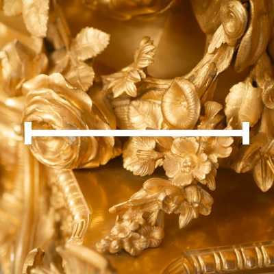 Obraz przedstawia złocone, bogato zdobione ornamenty w formie kwiatów i liści. W centralnej części widoczne są róże oraz inne drobne kwiaty i liście, które tworzą dekoracyjną kompozycję. Na pierwszym planie znajduje się biały pasek z dwoma pionowymi zakończeniami po bokach.